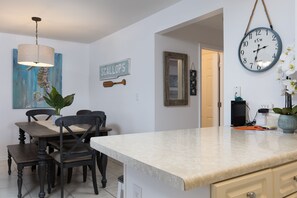 Un comedor con encimera de mármol, sillas de madera, lámpara colgante, un cartel que dice 'SCALLOPS', un espejo y un reloj de pared.