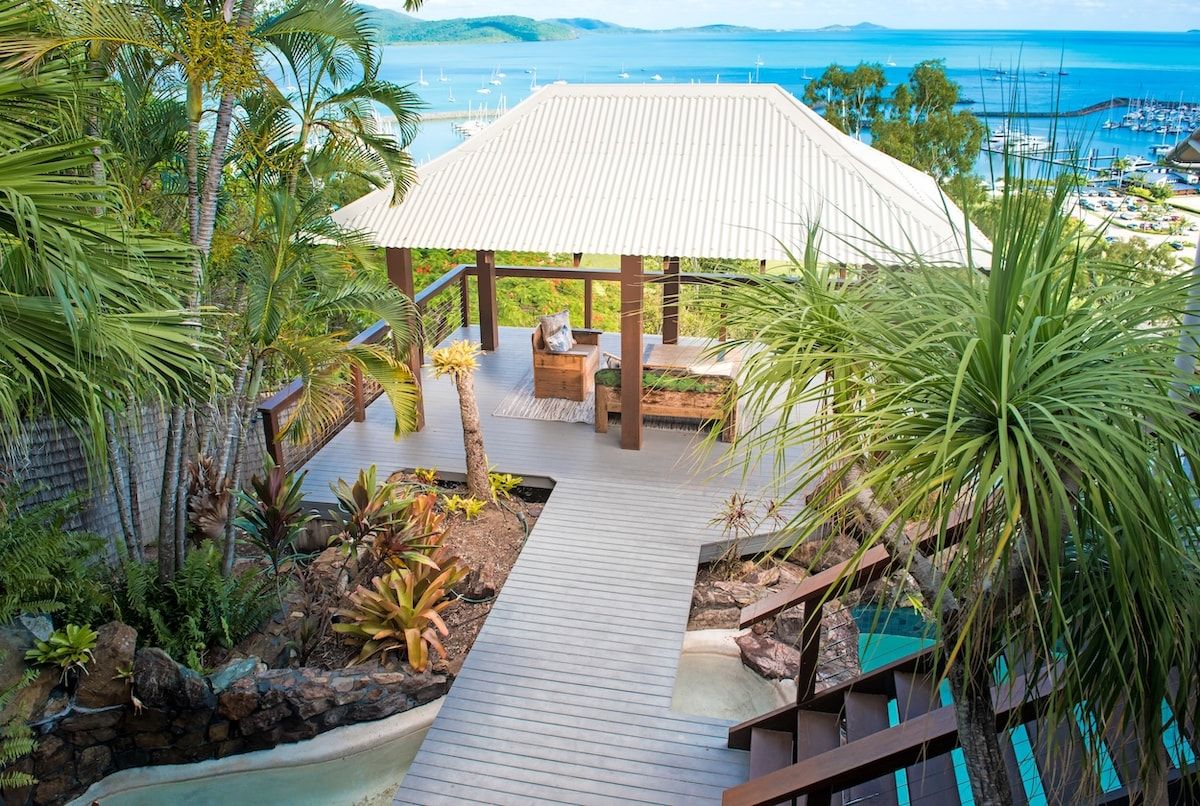 Un espace extérieur abrité, doté d’une terrasse en bois, entouré de plantes tropicales et donnant accès à une piscine.