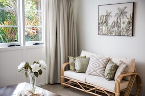 Ein Zimmer mit einem Korbsessel, einer Blumenvase, einem gerahmten Pflanzenmotiv an der Wand und einem Fenster mit Vorhängen.