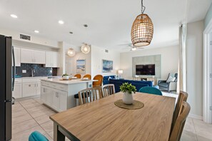 Uma cozinha moderna com armários brancos, uma sala de jantar com mesa de madeira e uma sala de estar com sofá azul e televisão.