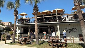 A restaurant with outdoor seating, palm trees, and a sign reading 'Sandos Beachside.' - Pet-Friendly Maravilla Condo–Beach Ramp, Pools, Free Beach Setup, 1BDR + Bunks (Miramar Beach)