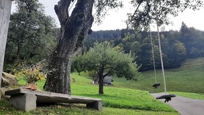 A rustic outdoor setting with a swing, a stone bench, and a small wooden structure in a grassy area with trees and hills in the background. - Vacation home with conservatory, fireplace and garden barbecue in a secluded location in the mountains (Tschagguns)