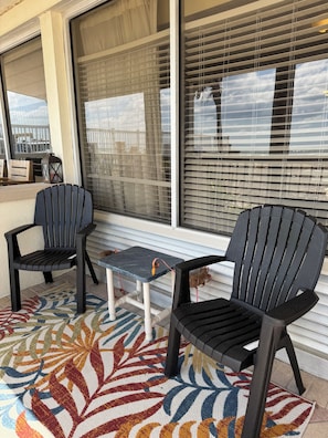 Eine Terrasse mit zwei Stühlen, einem kleinen Tisch, einem gemusterten Teppich und großen Fenstern mit Jalousien.