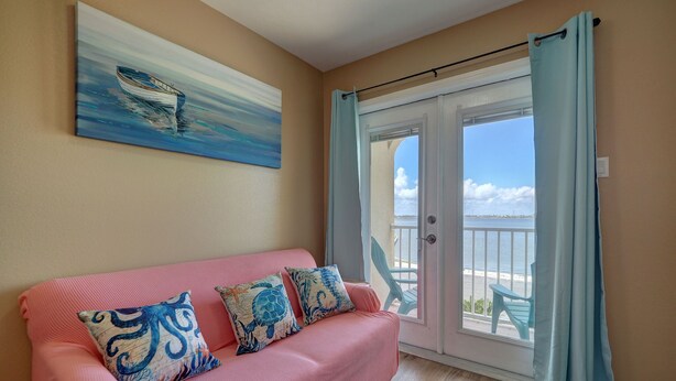 Une pièce avec un canapé rose, des coussins décoratifs, un tableau représentant un bateau et une vue sur la mer, visible à travers une porte vitrée.