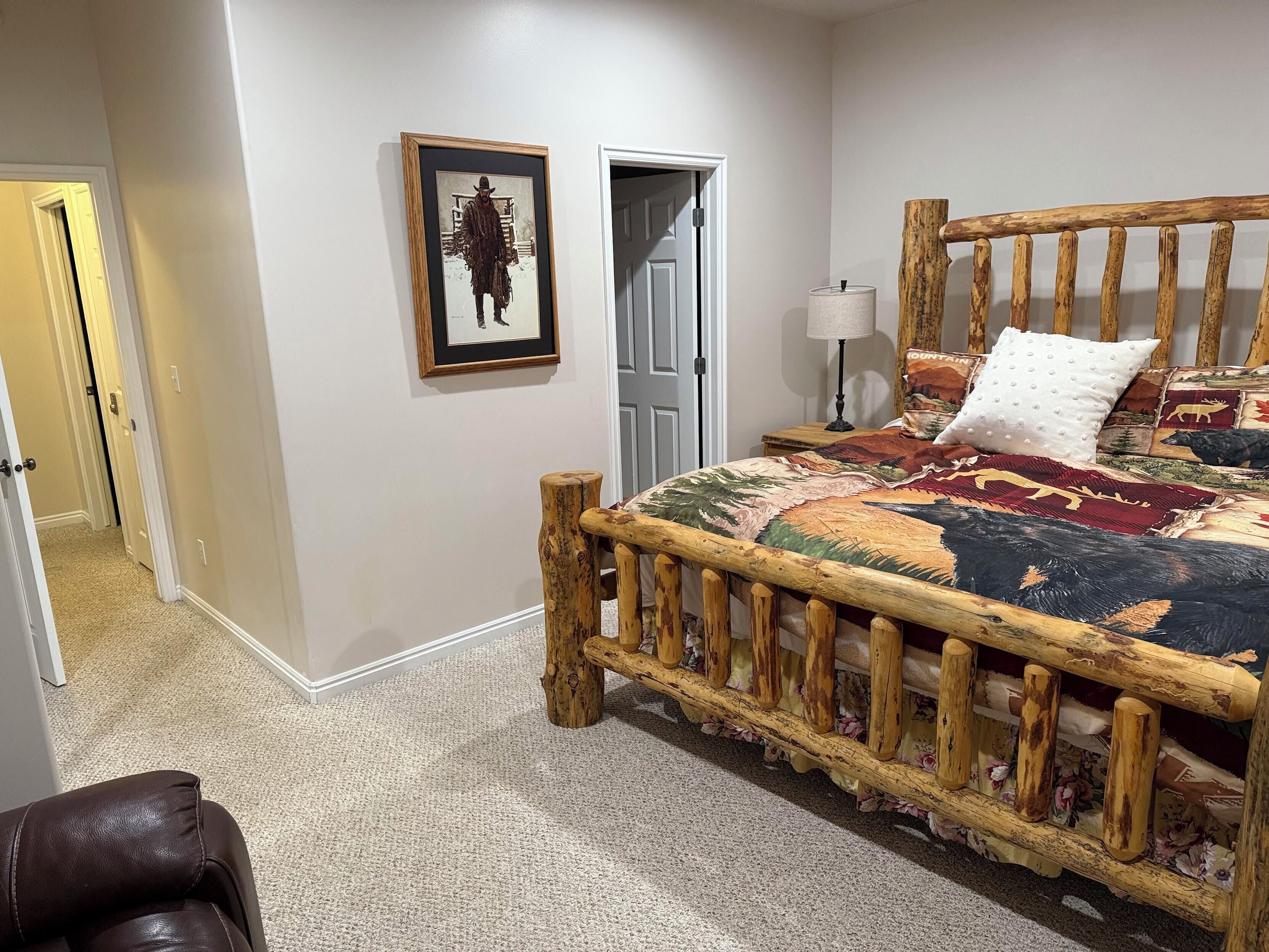 A bedroom with a wooden bed frame, a framed picture on the wall, a door, and a nightstand with a lamp.