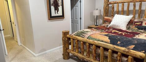 A bedroom with a wooden bed frame, a framed picture on the wall, a door, and a nightstand with a lamp.