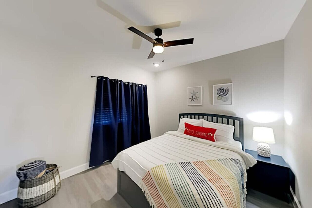 A bedroom with a bed, a ceiling fan, a nightstand with a lamp, and a window with curtains.