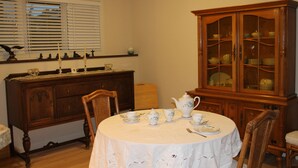 Een eetkamer met een ronde tafel gedekt voor thee, een houten kast met glazen deuren en een raam met jaloezieën.