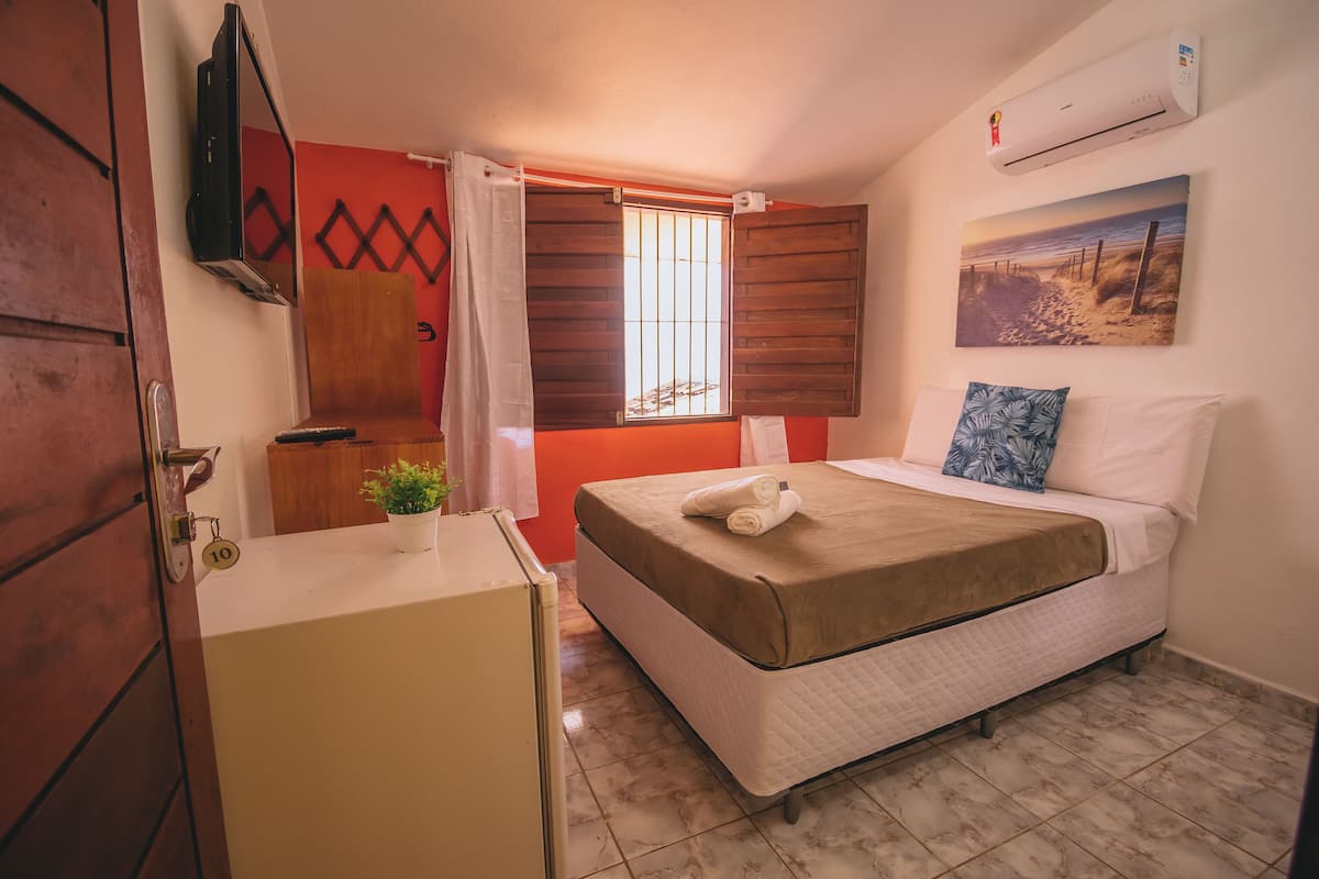 Un dormitorio con una cama, una ventana con contraventanas de madera, un aire acondicionado de pared y un cuadro de una playa.