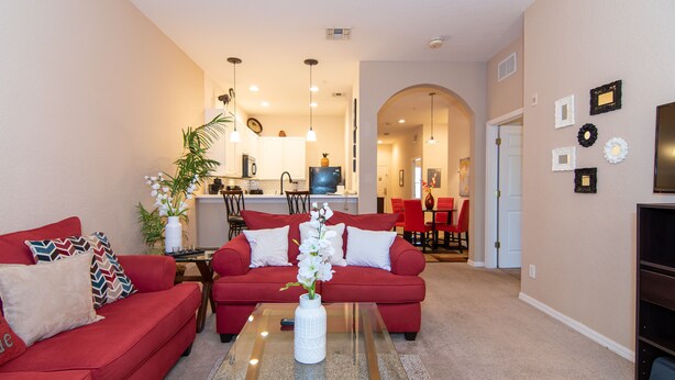 A living room with a red sofa, a coffee table with a vase, and a kitchen in the background.
