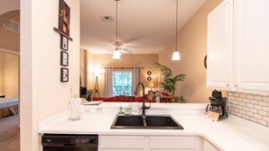 A kitchen with a dishwasher, double sink, and white cabinets. There is a ceiling fan, pendant lights, and a window with curtains.