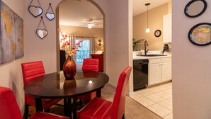 A dining area with a round table, red chairs, and a vase with flowers. There is a kitchen in the background with white cabinets and a black dishwasher.