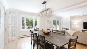 A dining room with a wooden table, six chairs, a chandelier, and a large window.