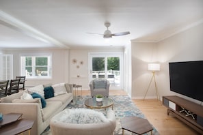 A spacious living room with a large TV, a ceiling fan, a sofa, a coffee table, and a dining area with chairs and a table. - Hudson Valley Hideaway - Heated Pool, Lake Access & Fenced Yard - Sleeps 8 (Copake)