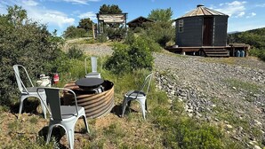Un área de estar rústica al aire libre con sillas de metal y una mesa de madera, una cabaña pequeña con techo de metal, y una estructura cubierta con una mesa y sillas al fondo.