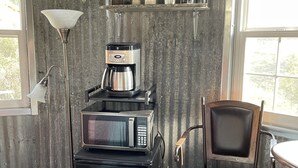 Cocina compacta con cafetera, microondas y refrigerador. Hay una silla y una lámpara cerca de la ventana.