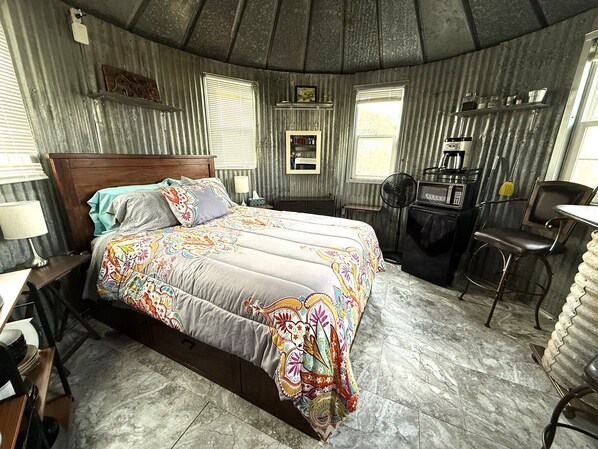 A room with a bed, a desk, a chair, a fan, and a coffee machine.