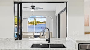 A modern kitchen with a built-in sink, a ceiling fan, and a view of a pool and greenery. - Seabreeze Oasis - 4 Br 3 Ba Lakefront Home w/ Heated Pool, Kayaks + more (Cape Coral)