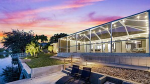 A modern house with a glass facade, outdoor seating, and a view of a body of water. - Seabreeze Oasis - 4 Br 3 Ba Lakefront Home w/ Heated Pool, Kayaks + more (Cape Coral)