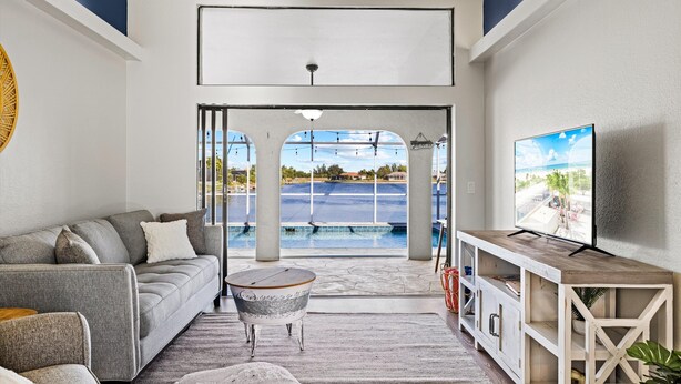 A modern living room with a grey sofa, a round coffee table, and a TV on a wooden stand. - Seabreeze Oasis - 4 Br 3 Ba Lakefront Home w/ Heated Pool, Kayaks + more (Cape Coral)