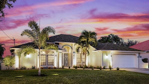 A modern house with a large garage, a well-maintained lawn, and a sunset sky. - Seabreeze Oasis - 4 Br 3 Ba Lakefront Home w/ Heated Pool, Kayaks + more (Cape Coral)