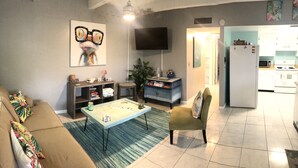 A living room with a sofa, coffee table, and a TV. There's a kitchen area with a fridge and microwave. - Flirt with the Flamingos in Beach Bliss ~Just Steps to the Sand (FORT PIERCE)