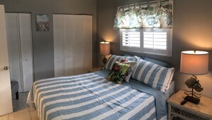 A bedroom with a bed, two bedside tables, a window with curtains, a wardrobe, and a wall with a painting. - Flirt with the Flamingos in Beach Bliss ~Just Steps to the Sand (FORT PIERCE)