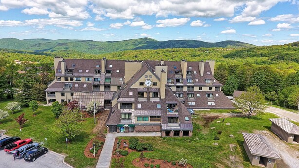A multi-story hotel with a central clock tower, surrounded by lush greenery and mountains in the background.