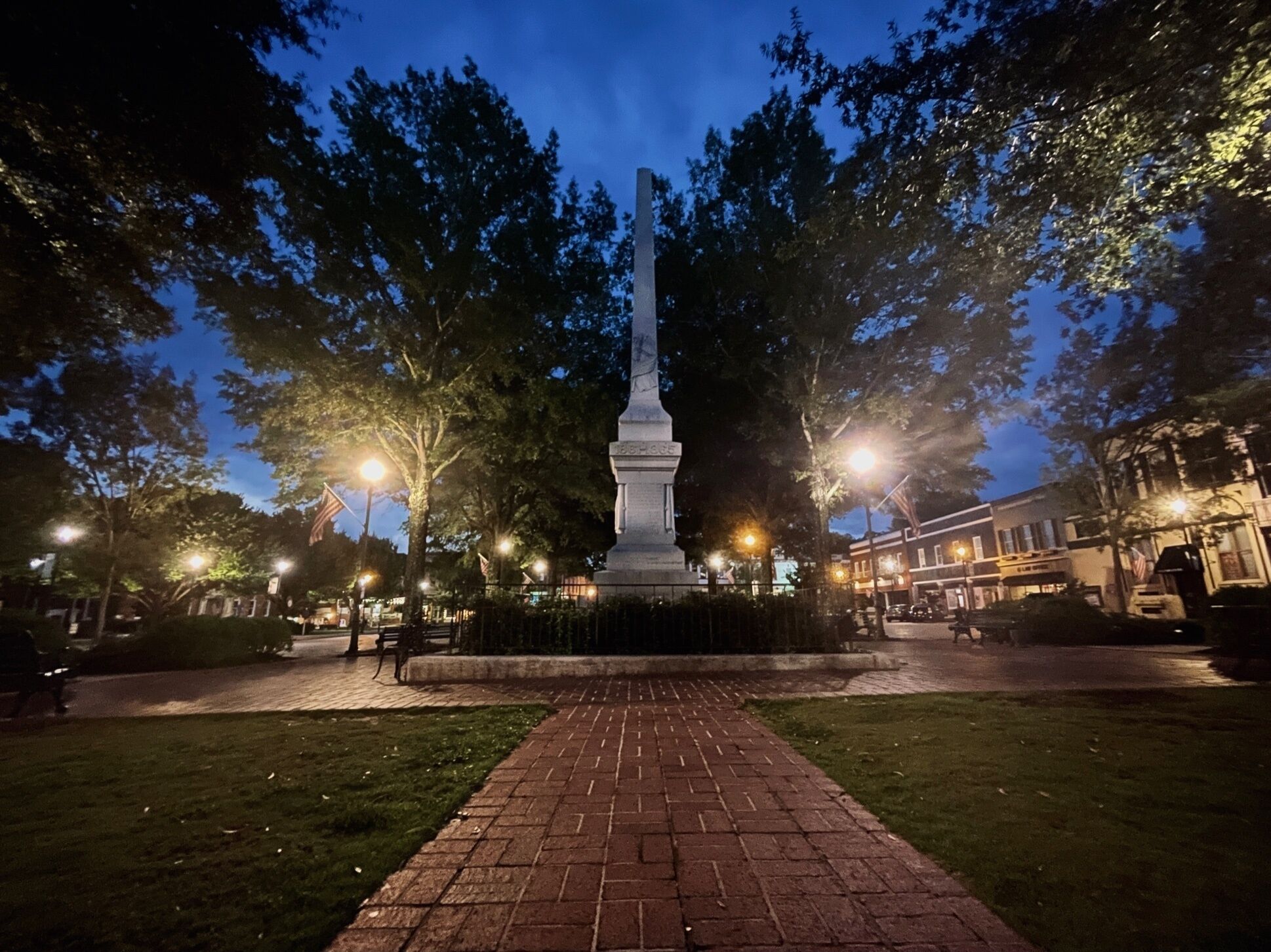 A monument in a park with a brick pathway leading to it, surrounded by trees and street lamps.