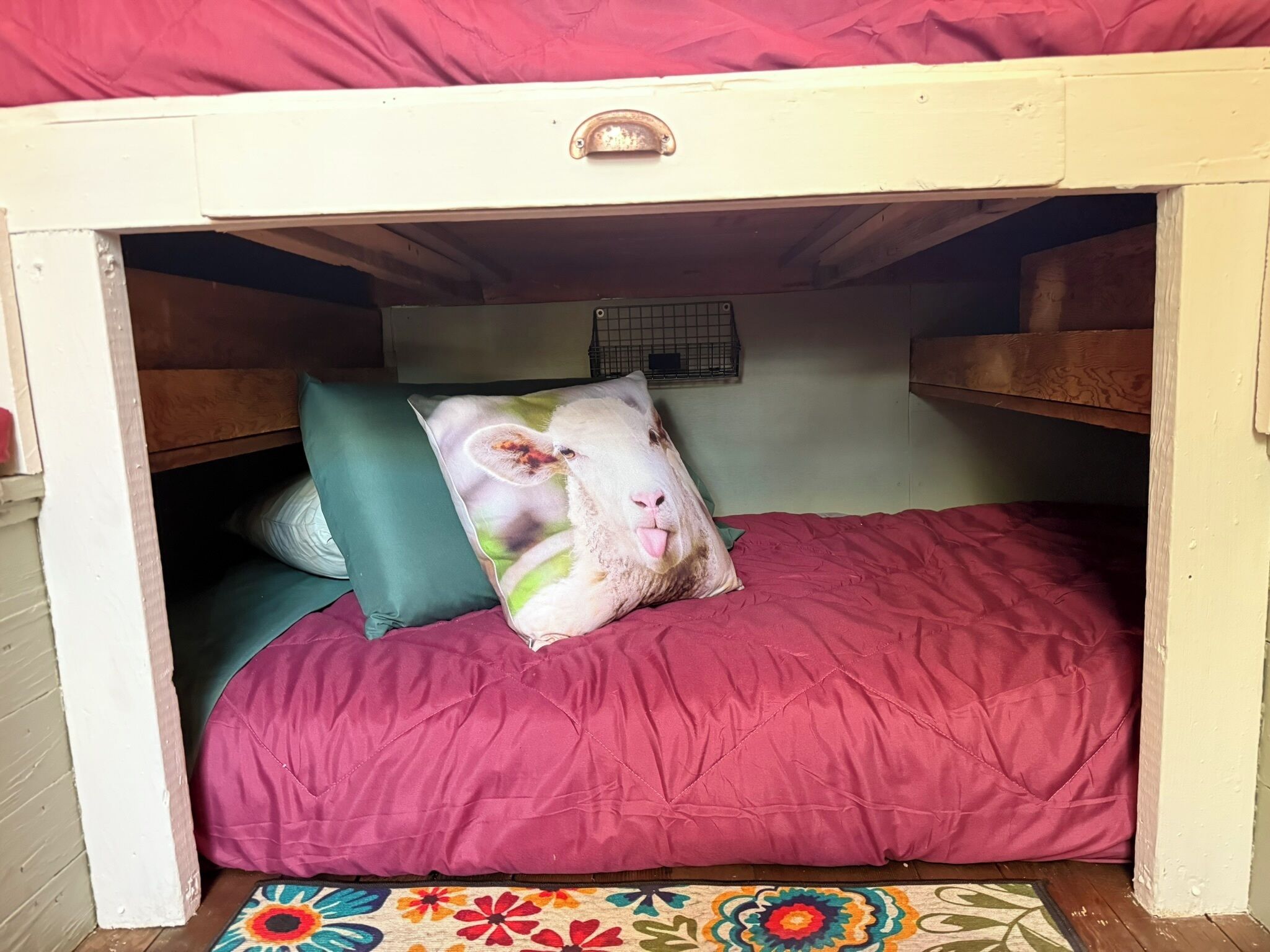 Ein kleines Bett mit einer roten Matratze und einem farbenfrohen Blumenteppich am Fußende.