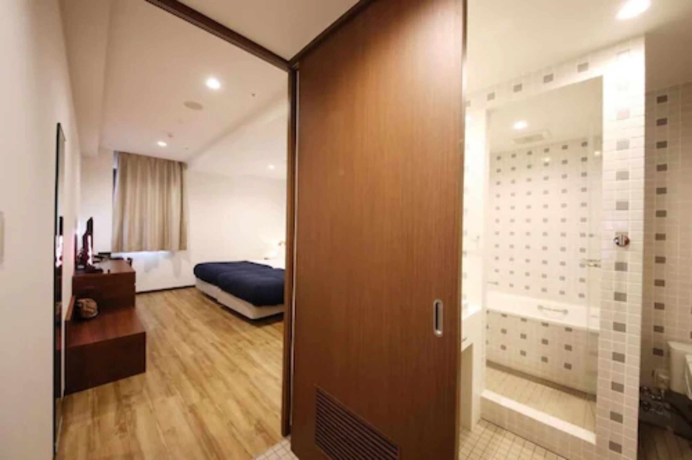 Habitación de hotel con piso de madera, cama, escritorio con silla y zona de ducha con paredes de azulejos.