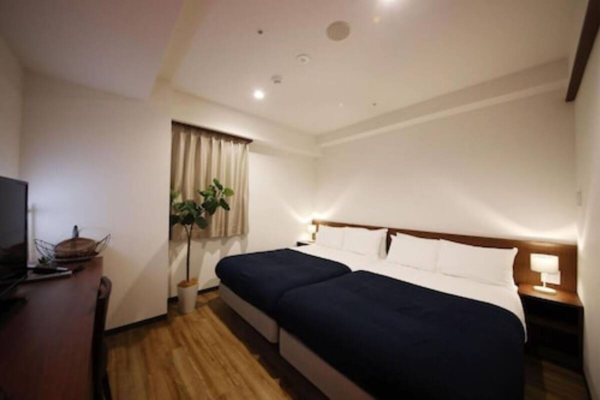 A hotel room with a wooden floor, a bed with dark bedding, a bedside table with a lamp, a TV on a stand, and a potted plant near the window.