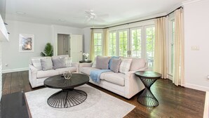 Un salon comprenant un canapé d’angle, une table basse et une table d’appoint. On y trouve également un ventilateur de plafond, une fenêtre avec des rideaux et une plante.