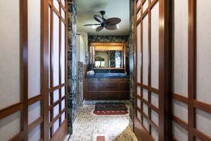 A narrow hallway with a wooden floor, a tiled floor mat, a ceiling fan, and a bathroom with a sink and mirror.