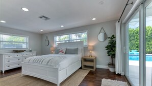 Camera da letto con un letto grande, un comodino, una lampada, una pianta in vaso e vista su una piscina attraverso una porta scorrevole in vetro.