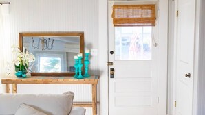Una habitación con una puerta blanca, una consola de madera con candelabros turquesa, un espejo y un florero con flores blancas.