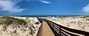 En træpromenade der fører til en strand med klitter og en klar himmel.