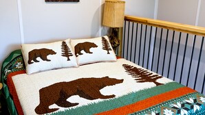 Uma cama com um edredom temático de urso, uma pintura de um incêndio florestal e um abajur em um corrimão de madeira.