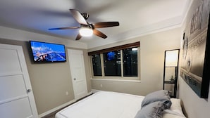 Une chambre à coucher avec un lit, un ventilateur de plafond, un téléviseur fixé au mur, une fenêtre et une table de chevet avec une lampe.