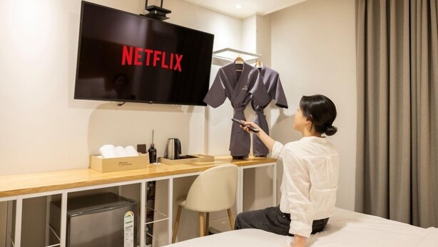 A hotel room with a bed, a desk with a TV displaying 'NETFLIX', a minibar, and a wardrobe with clothes. - Cheongju Hotel Dictionary (Cheongju)