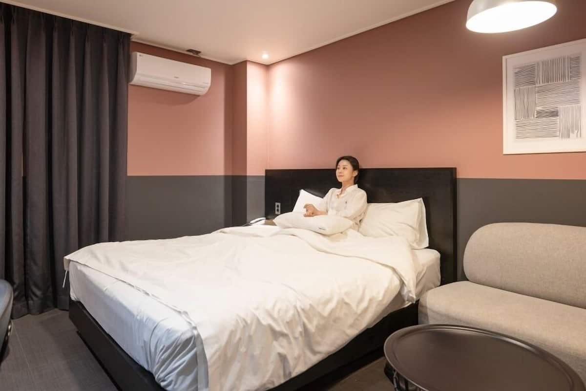Habitación de hotel con cama, una silla, un sofá, un equipo de aire acondicionado y una obra de arte en la pared.