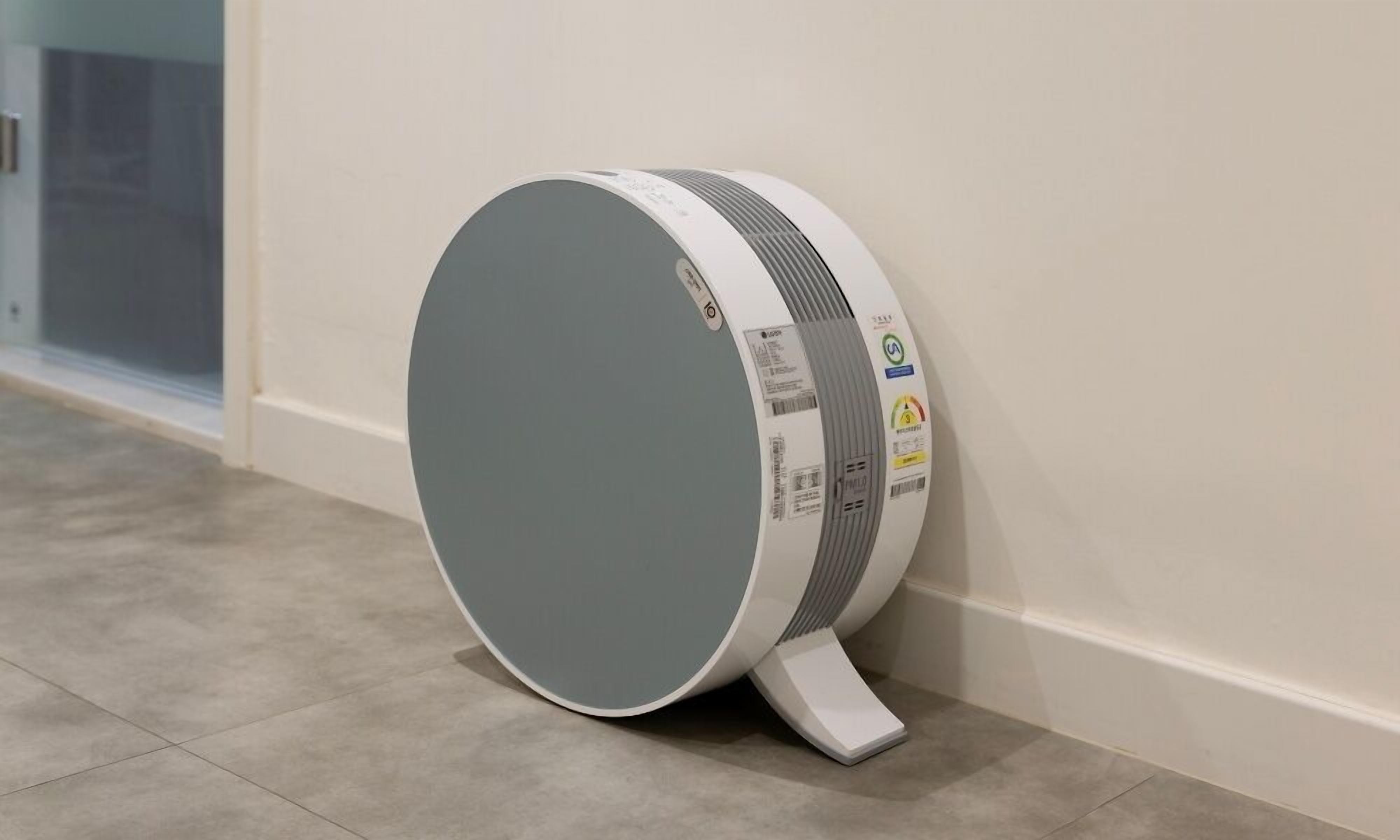 A modern, cylindrical air purifier with a grey top and white base, placed against a white wall in a hallway with tiled flooring.