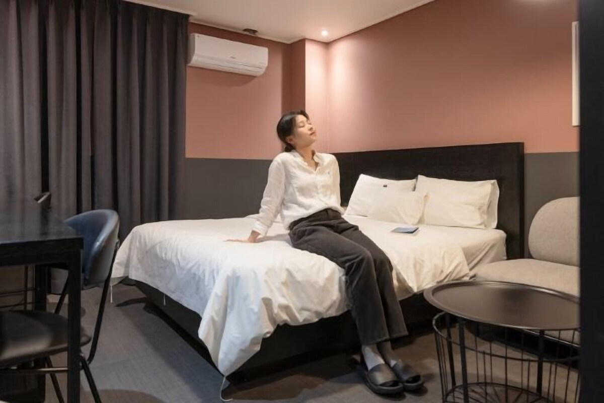 A hotel room with a bed, a small table, a chair, and an air conditioning unit on the wall.