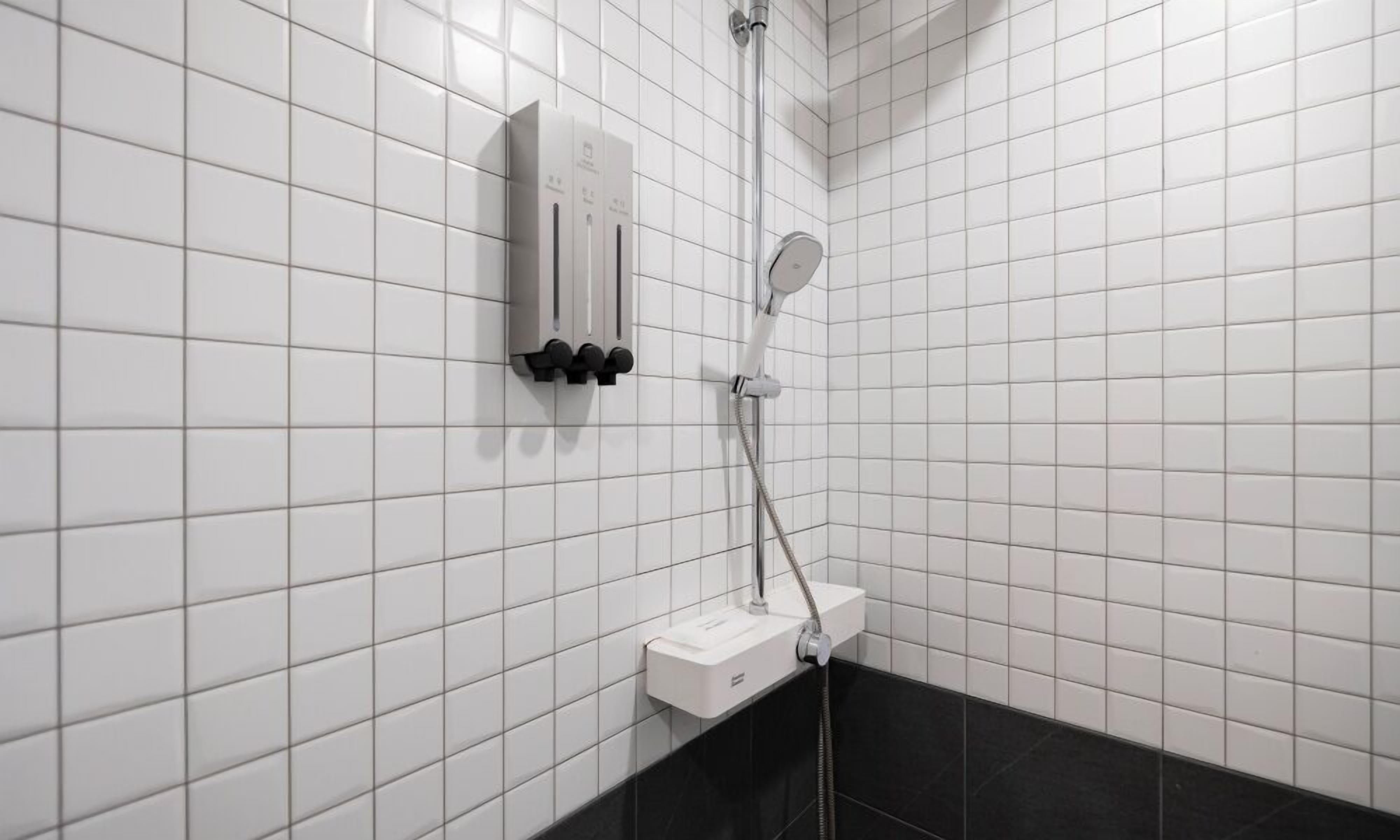 A bathroom with white tiled walls, a black tiled floor, a shower with a handheld showerhead, and a control panel with multiple buttons.