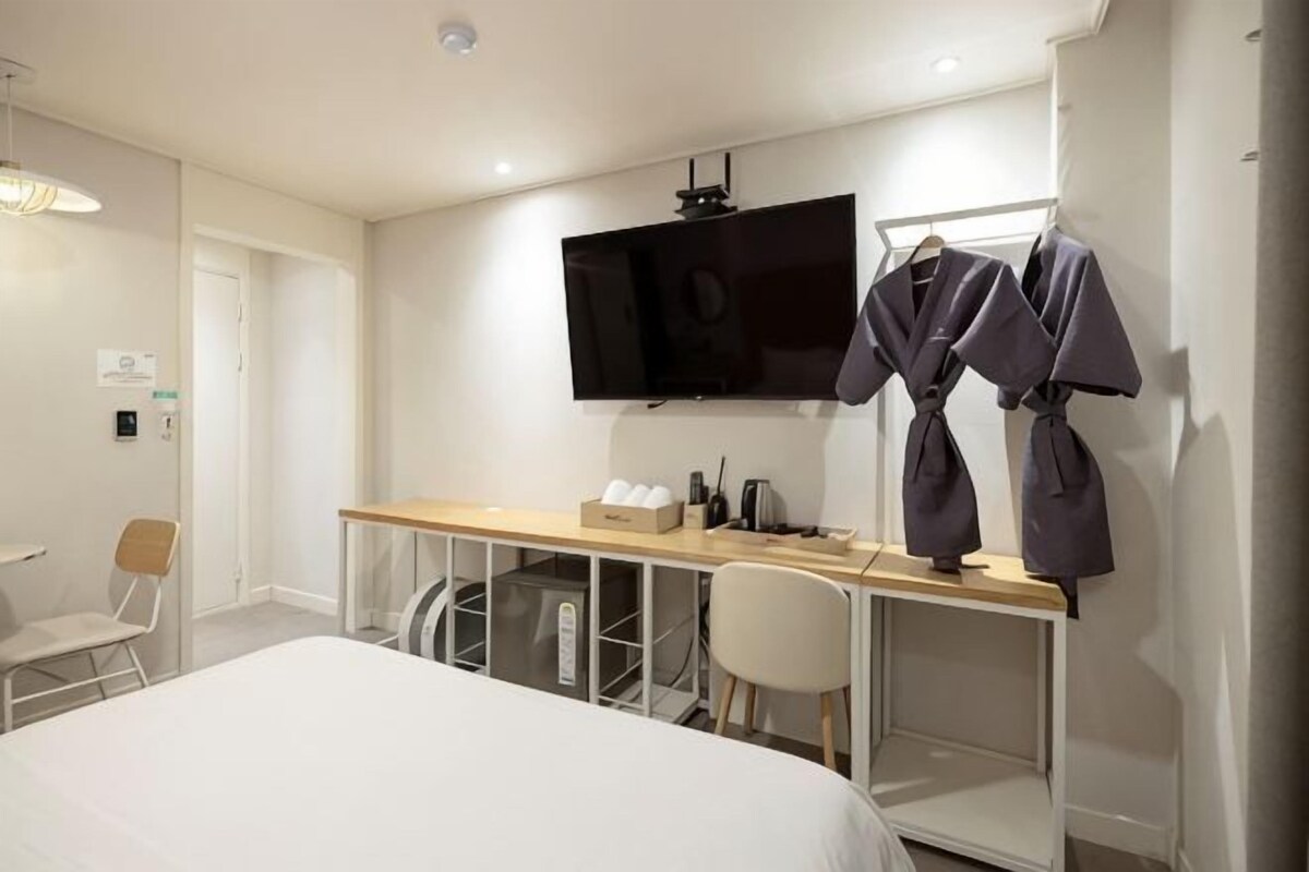 A hotel room with a bed, a desk with a flat-screen TV, a chair, and a wardrobe with hanging robes.