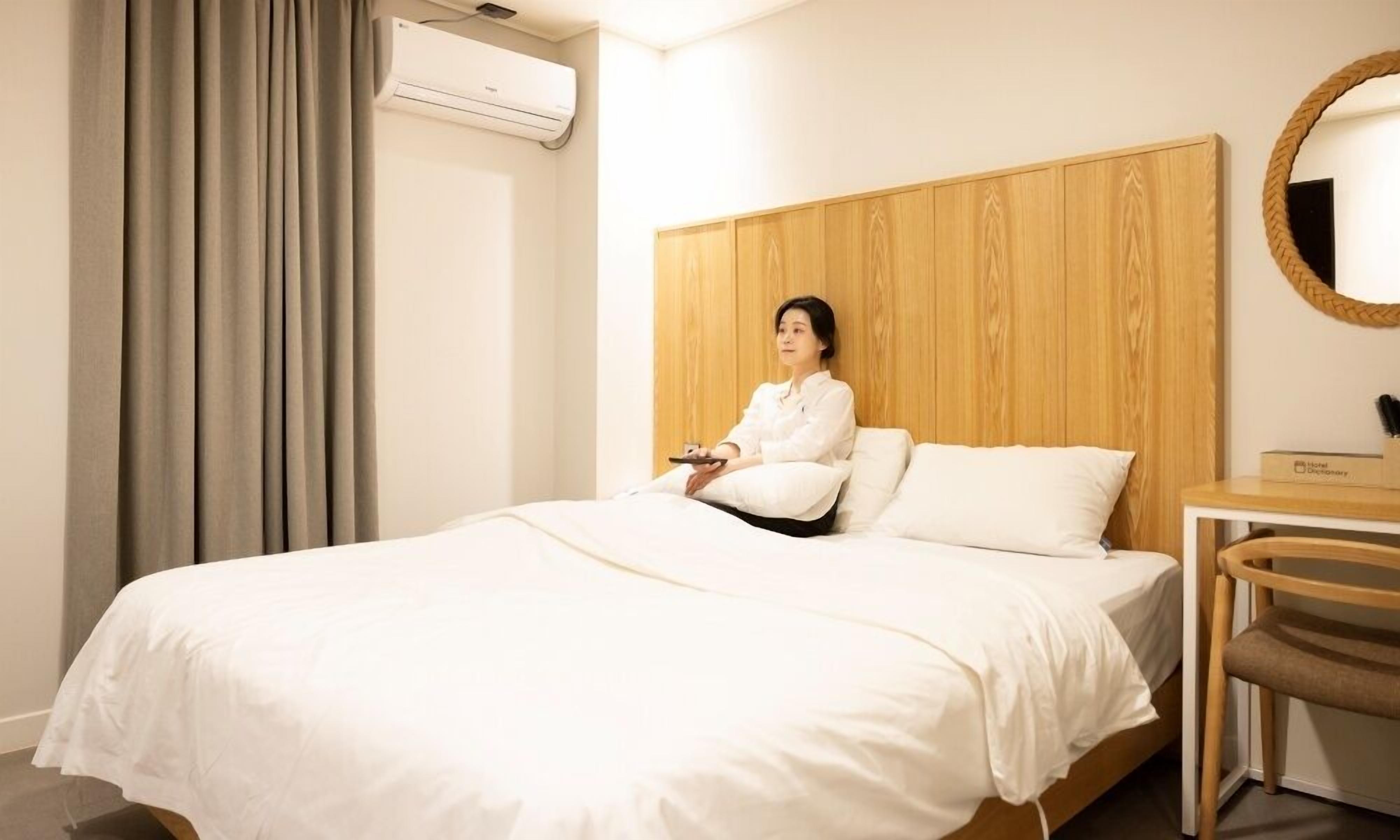 A hotel room with a wooden headboard, a bed with white bedding, a bedside table with a lamp, and an air conditioning unit.