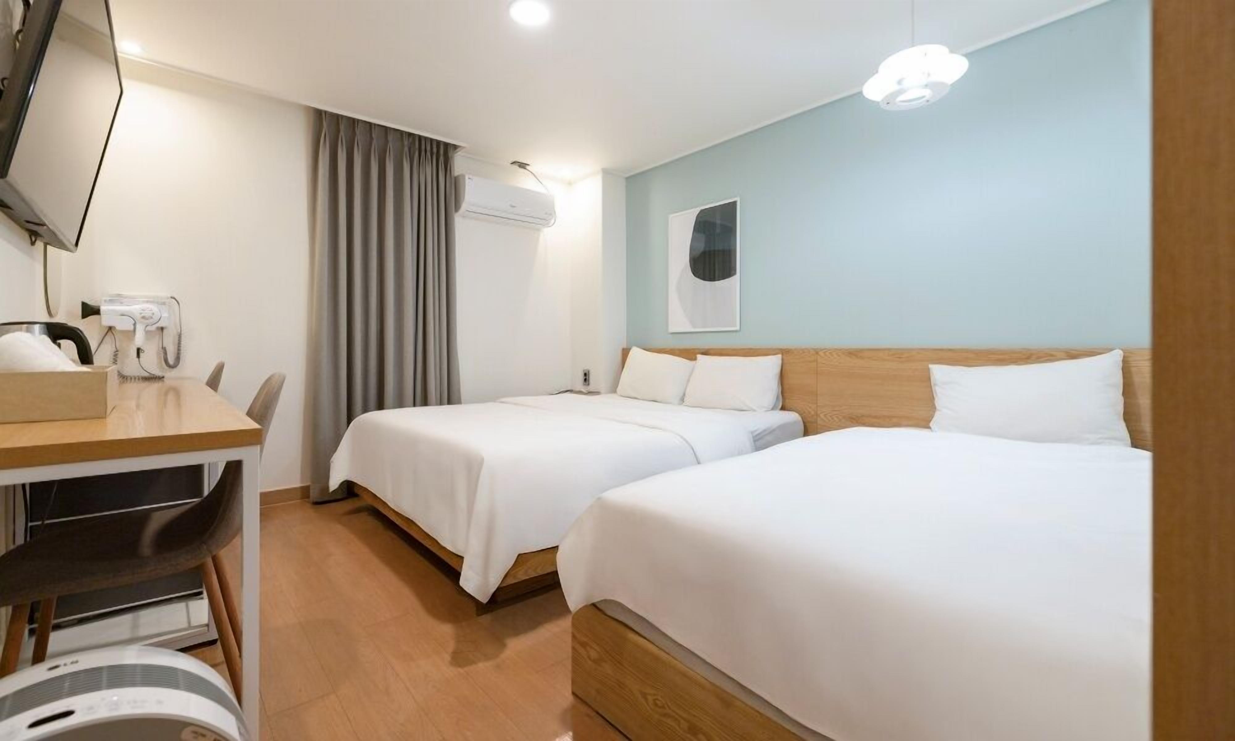 A hotel room with two beds, a desk with a chair, a hairdryer, and an air conditioning unit.