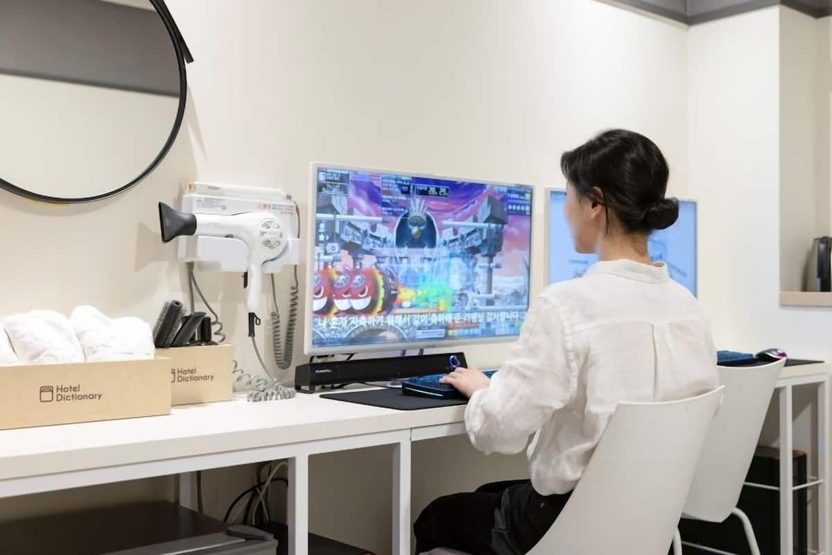 Una persona sentada en un escritorio con una computadora, un secador de pelo y una caja etiquetada 'Hotel Dictionary'.