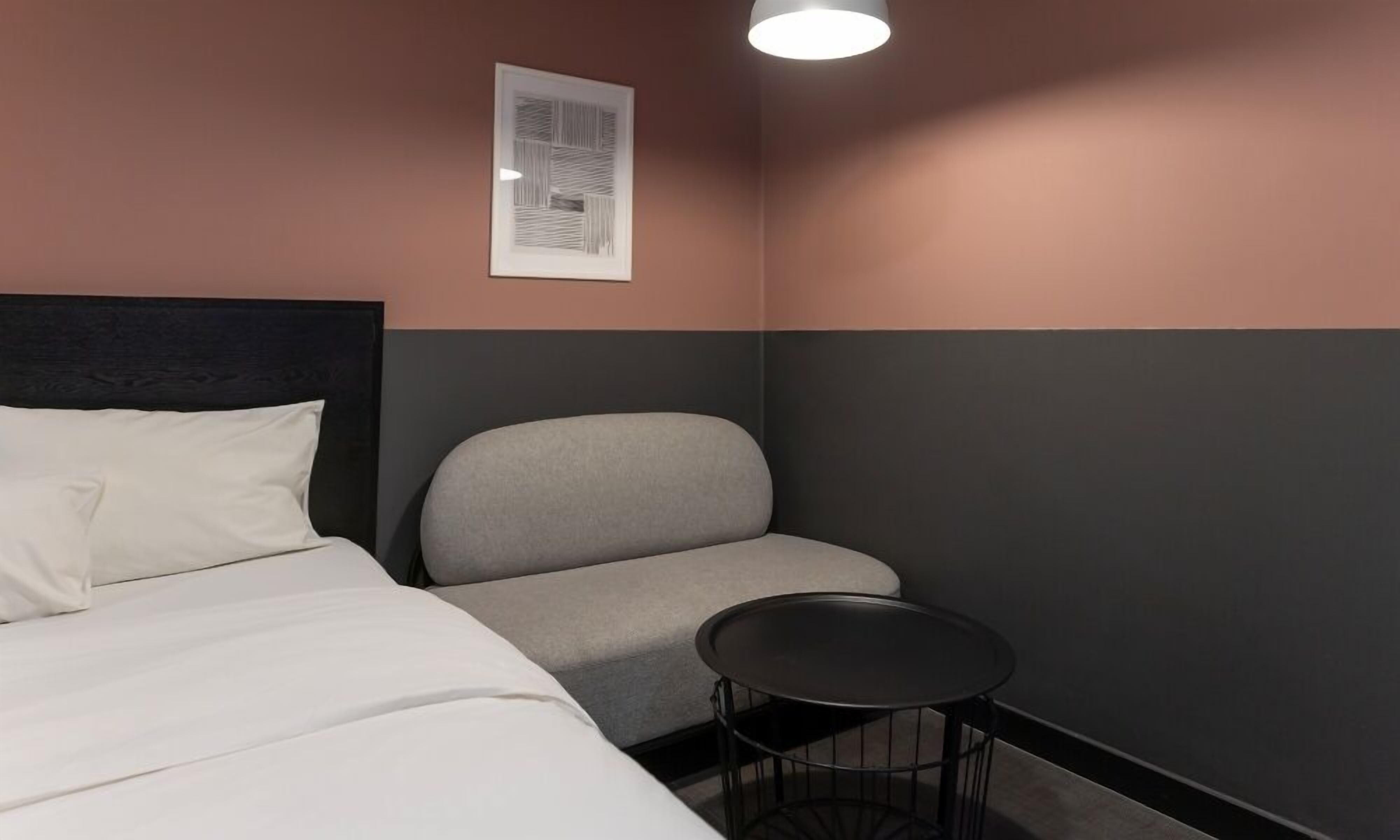 A hotel room with a bed, a grey armchair, a small round table, and a framed picture on the wall.