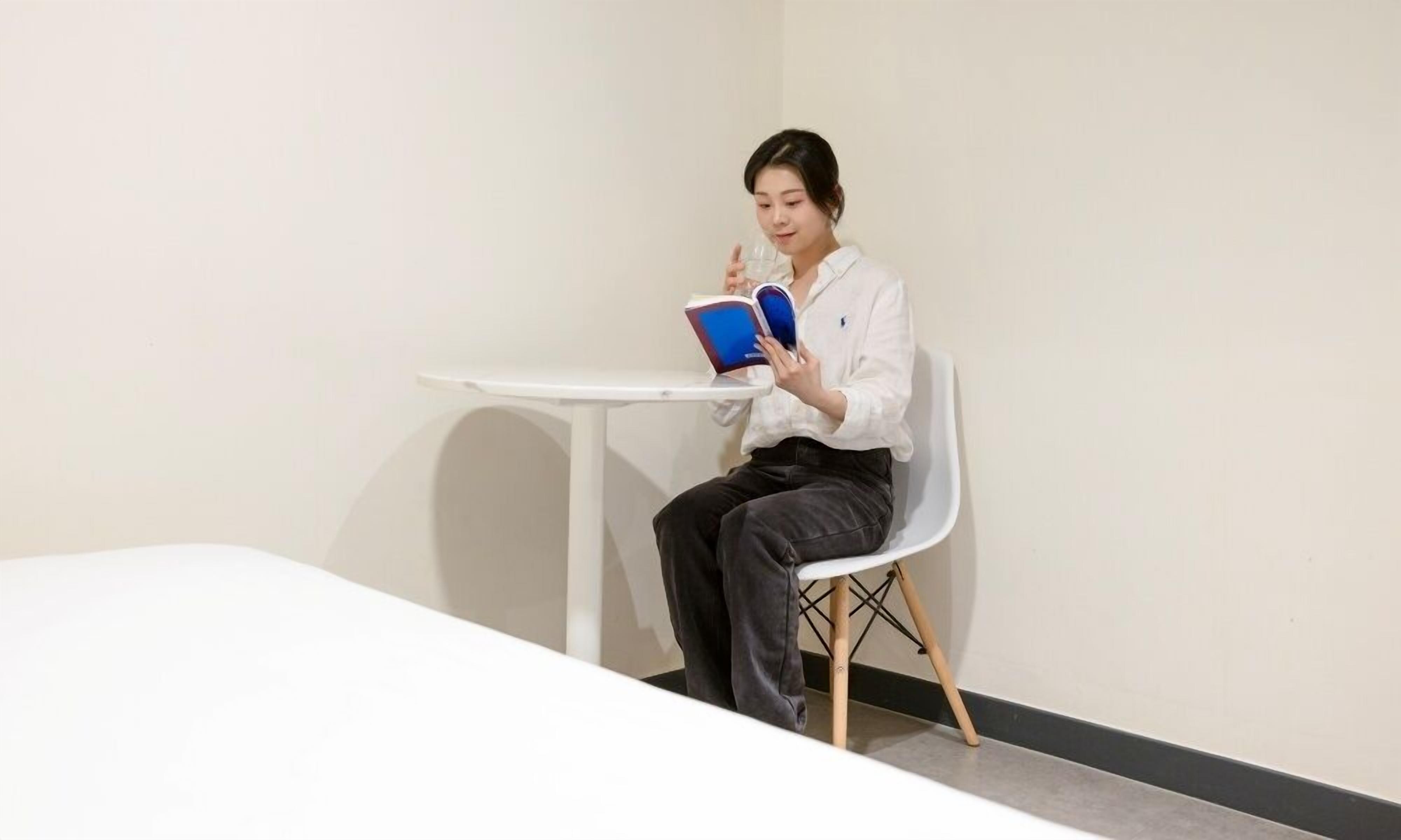 A person seated at a small white table with a book, in a room with plain walls and a simple floor.